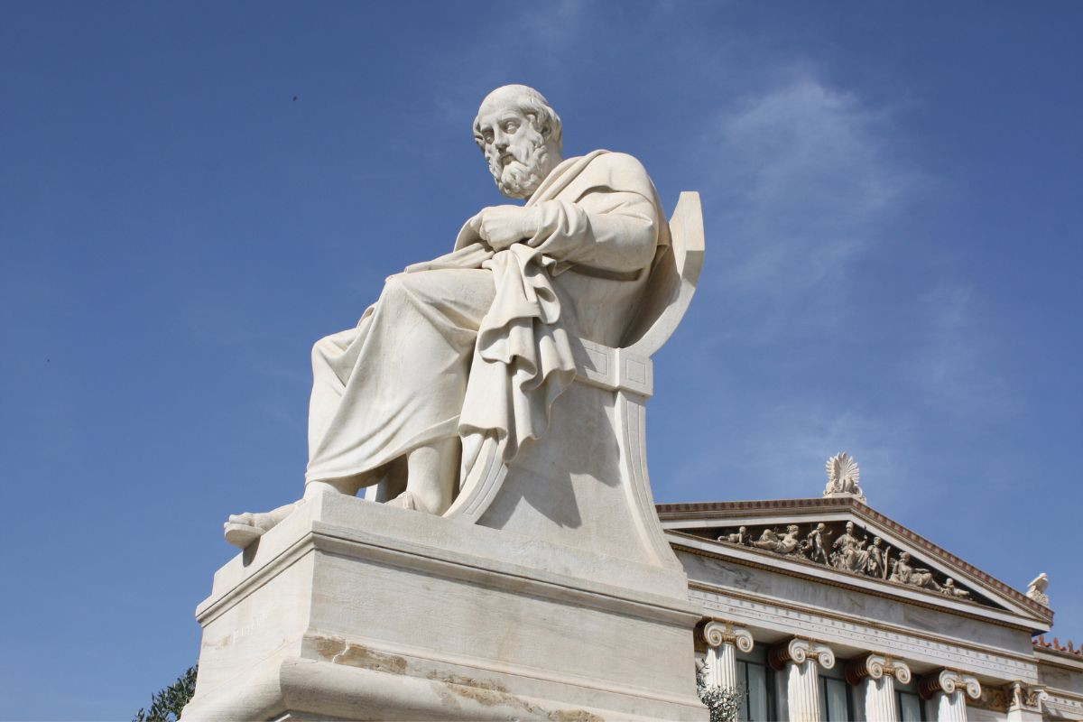 The image shows a statue of the ancient Greek philosopher Plato, seated and draped in robes, with a contemplative expression. The statue is placed in front of a classical building with columns and ornate decorations, under a clear blue sky.